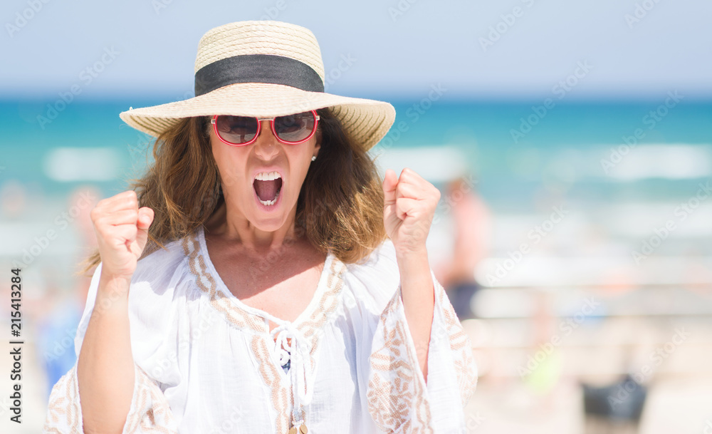 Middle age brunette woman wearing summer hat on vacations by the beach annoyed and frustrated shouting with anger, crazy and yelling with raised hand, anger concept