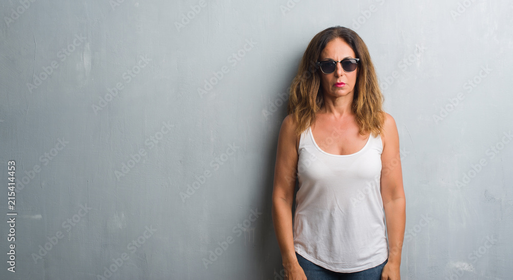 Middle age hispanic woman over grey wall wearing sunglasses with serious expression on face. Simple and natural looking at the camera. Middle age hispanic woman over grey wall wearing sunglasses with serious expression on face. Simple and natural looking at the camera.