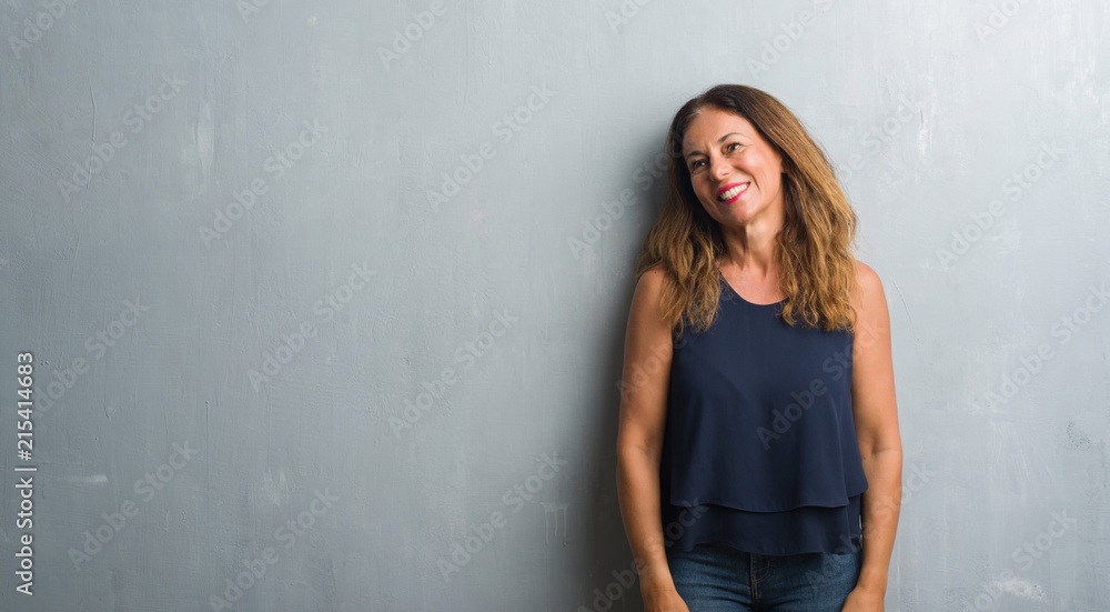 Middle age hispanic woman standing over grey grunge wall looking away to side with smile on face, natural expression. Laughing confident. Middle age hispanic woman standing over grey grunge wall looking away to side with smile on face, natural expression. Laughing confident.