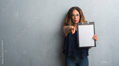 Middle age hispanic woman standing over grey grunge wall holding clipboard pointing with finger to the camera and to you, hand sign, positive and confident gesture from the front