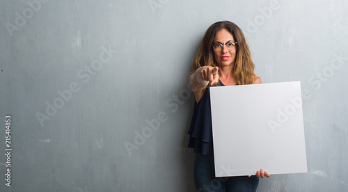 Middle age hispanic woman standing over grey grunge wall holding blank banner pointing with finger to the camera and to you, hand sign, positive and confident gesture from the front