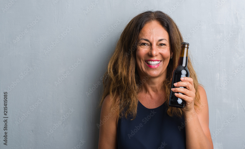 Middle age hispanic woman standing over grey grunge wall holding beer bottle with a happy face standing and smiling with a confident smile showing teeth Middle age hispanic woman standing over grey grunge wall holding beer bottle with a happy face standing and smiling with a confident smile showing teeth