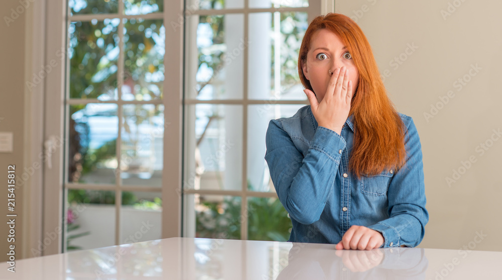 Fototapeta premium Redhead woman at home cover mouth with hand shocked with shame for mistake, expression of fear, scared in silence, secret concept