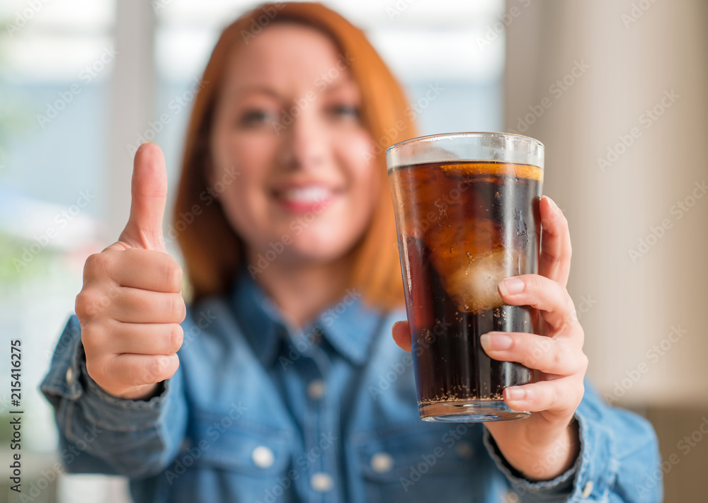 Redhead woman holding soda refreshment happy with big smile doing ok sign, thumb up with fingers, excellent sign Redhead woman holding soda refreshment happy with big smile doing ok sign, thumb up with fingers, excellent sign