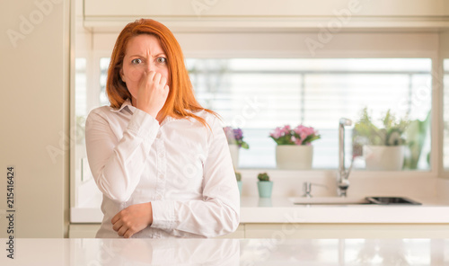 Redhead woman at kitchen smelling something stinky and disgusting, intolerable smell, holding breath with fingers on nose. Bad smells concept.