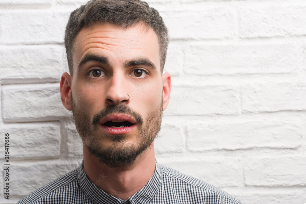 Young adult man standing over white brick wall scared in shock with a surprise face, afraid and excited with fear expression Young adult man standing over white brick wall scared in shock with a surprise face, afraid and excited with fear expression