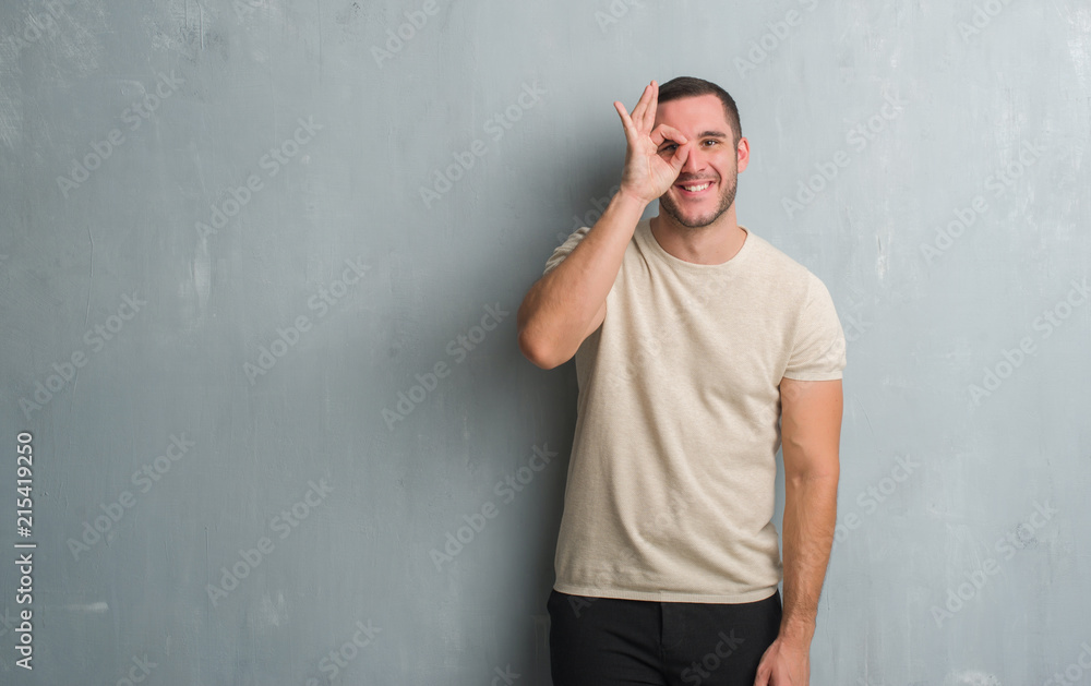 Young caucasian man over grey grunge wall doing ok gesture with hand smiling, eye looking through fingers with happy face.