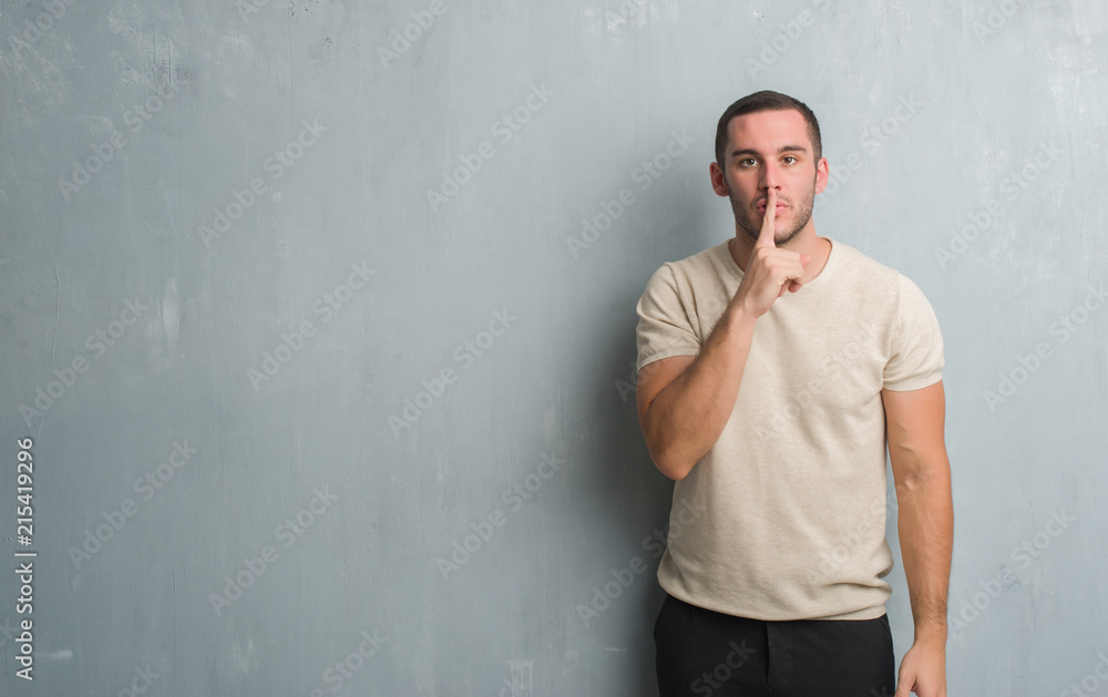 Young caucasian man over grey grunge wall asking to be quiet with finger on lips. Silence and secret concept.