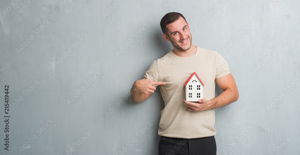 Young caucasian real state agent man over grey grunge wall holding house very happy pointing with hand and finger Young caucasian real state agent man over grey grunge wall holding house very happy pointing with hand and finger