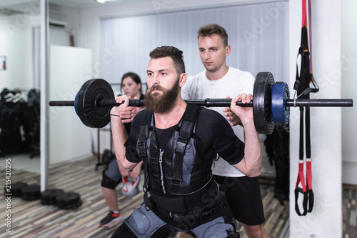 Young bearded man and woman doing exercises in electrical muscular stimulation suit with their personal trainer at rehabilitation center.
