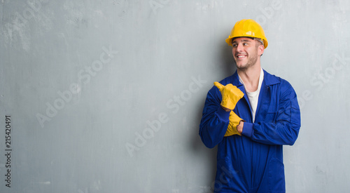 Young caucasian man over grey grunge wall wearing contractor uniform and safety helmet pointing and showing with thumb up to the side with happy face smiling
