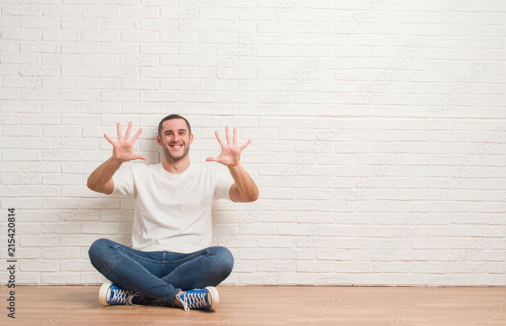 Young caucasian man sitting on the floor over white brick wall showing and pointing up with fingers number ten while smiling confident and happy. Young caucasian man sitting on the floor over white brick wall showing and pointing up with fingers number ten while smiling confident and happy.