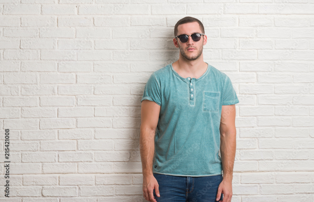 Young caucasian man standing over white brick wall wearing sunglasses with a confident expression on smart face thinking serious Young caucasian man standing over white brick wall wearing sunglasses with a confident expression on smart face thinking serious