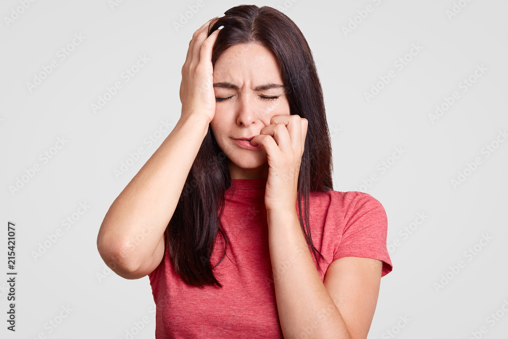 People and sadness concept. Stressed brunette female keeps eyes closed, touches head, feels pain, being in panic after divorce with husband, dressed in casual t shirt, isolated over white studio wall People and sadness concept. Stressed brunette female keeps eyes closed, touches head, feels pain, being in panic after divorce with husband, dressed in casual t shirt, isolated over white studio wall