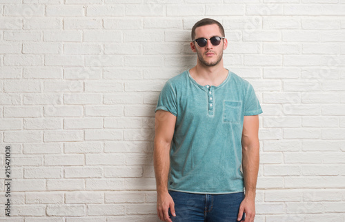 Young caucasian man standing over white brick wall wearing sunglasses with a confident expression on smart face thinking serious