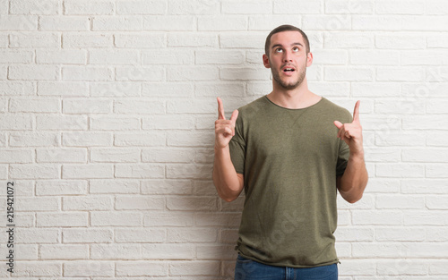 Young caucasian man standing over white brick wall amazed and surprised looking up and pointing with fingers and raised arms.