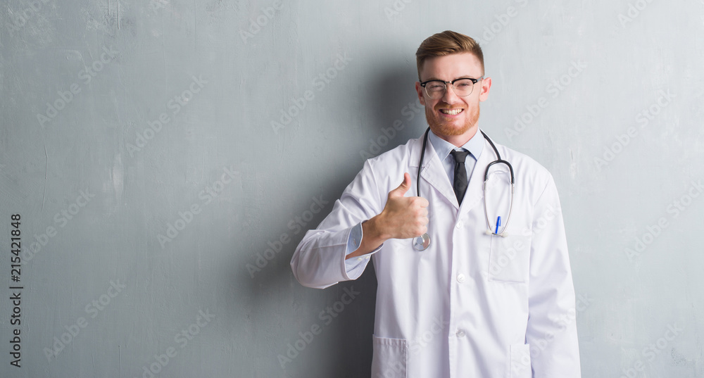 Young redhead doctor man over grey grunge wall happy with big smile doing ok sign, thumb up with fingers, excellent sign Young redhead doctor man over grey grunge wall happy with big smile doing ok sign, thumb up with fingers, excellent sign