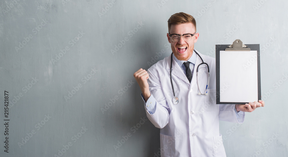 Young redhead doctor man over grey grunge wall holding clipboard screaming proud and celebrating victory and success very excited, cheering emotion Young redhead doctor man over grey grunge wall holding clipboard screaming proud and celebrating victory and success very excited, cheering emotion