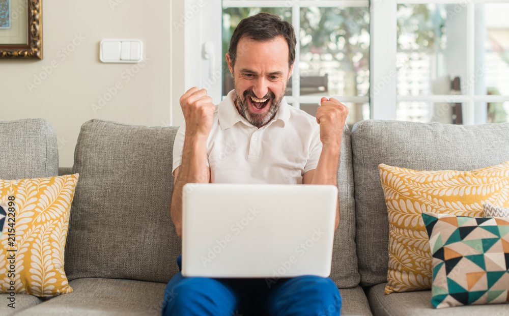 Middle age man using laptop at sofa screaming proud and celebrating victory and success very excited, cheering emotion Middle age man using laptop at sofa screaming proud and celebrating victory and success very excited, cheering emotion
