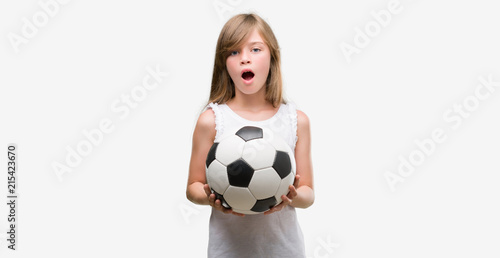Young blonde toddler holding football ball scared in shock with a surprise face  afraid and excited with fear expression