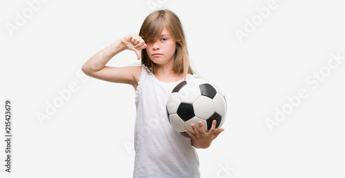 Young blonde toddler holding football ball with angry face, negative sign showing dislike with thumbs down, rejection concept