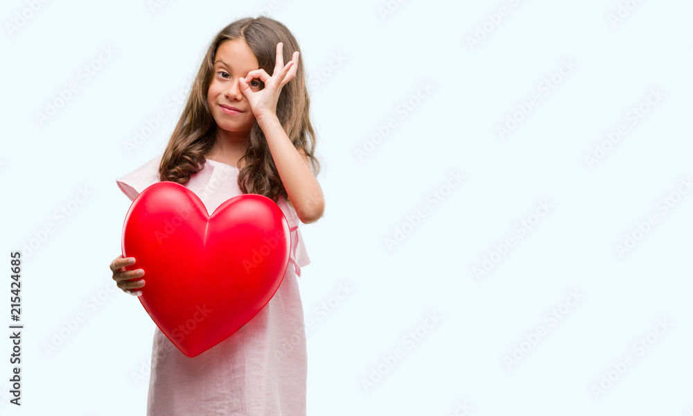 Brunette hispanic girl holding red heart with happy face smiling doing ok sign with hand on eye looking through fingers Brunette hispanic girl holding red heart with happy face smiling doing ok sign with hand on eye looking through fingers