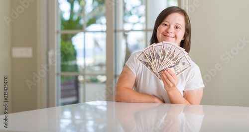 Down syndrome woman at home holding dollars with a happy face standing and smiling with a confident smile showing teeth