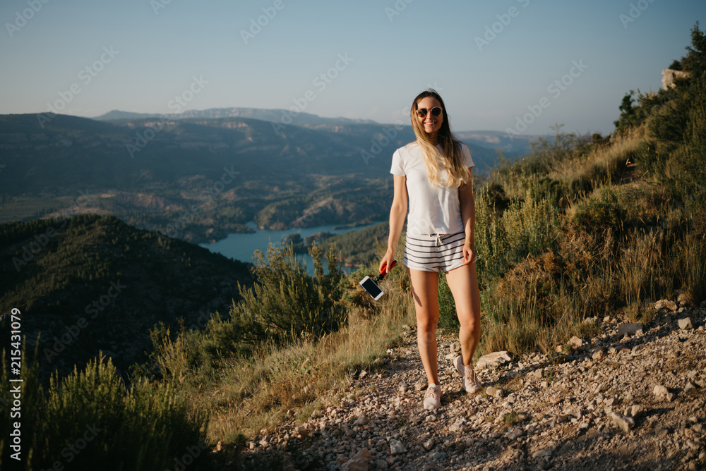 Fototapeta premium Pretty girl posing and smiling on the sandy road with canyon and emerald lake on the background on the sunset in Spain. Traveler in the mountains.