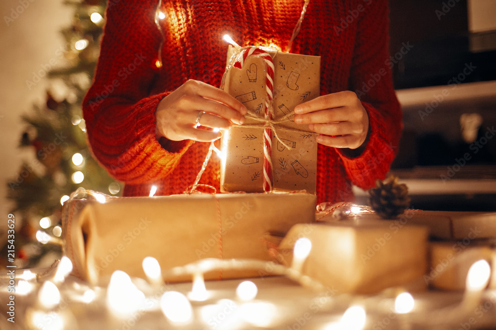 Fototapeta premium christmas magic moments. hands holding christmas present in lights in evening festive room under tree in illumination. space for text. girl opening modern craft gifts with candy cane