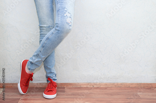 Woman fashion concept. Female legs in jeans and sneakers near the wall.