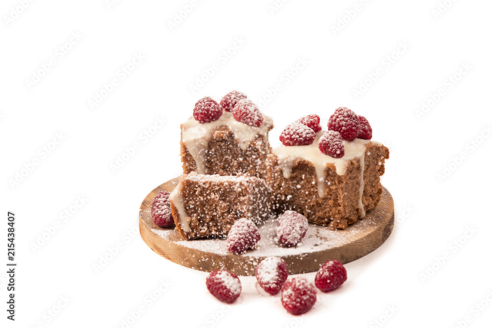 The chocolate cake decorated by white glaze and fresh berries of raspberry is strewed with icing sugar lies on a wooden round support. Separately on a white background. Horizontal shot. The place for The chocolate cake decorated by white glaze and fresh berries of raspberry is strewed with icing sugar lies on a wooden round support. Separately on a white background. Horizontal shot. The place for