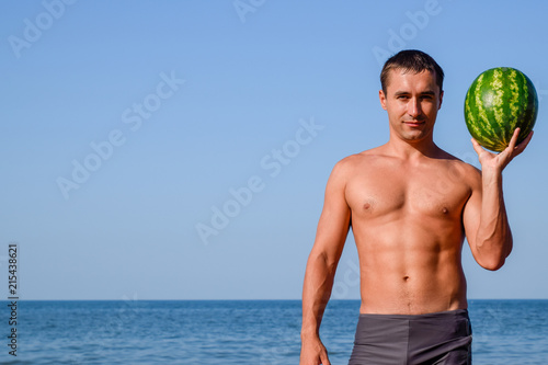 A sporty man is standing on the beach and holding a whole watermelon. A ripe watermelon in the hands of a man. photo