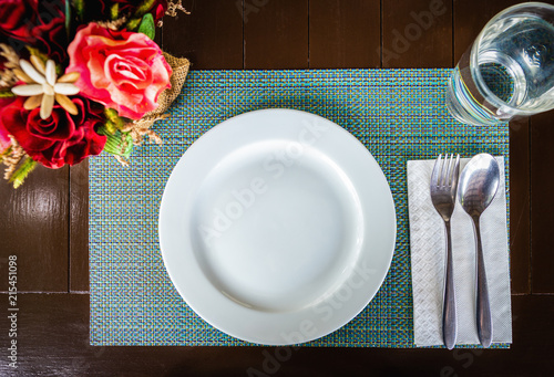 Top view of white ceramic dish with spoon and fork on tablemat on brown wooden table. Glass of water and sweet bouquet of flowers are beside. photo