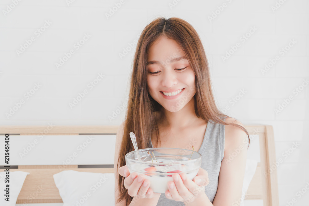 Asian beautiful smiling woman holds a glass bowl of yogurt and strawberry fruit on white bed in morning.Concept of women's health care. Asian beautiful smiling woman holds a glass bowl of yogurt and strawberry fruit on white bed in morning.Concept of women's health care.