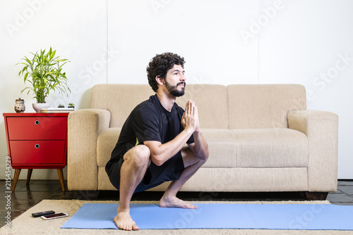 Beautiful caucasian bearded man in black clothes on blue yogamat doing garland pose photo