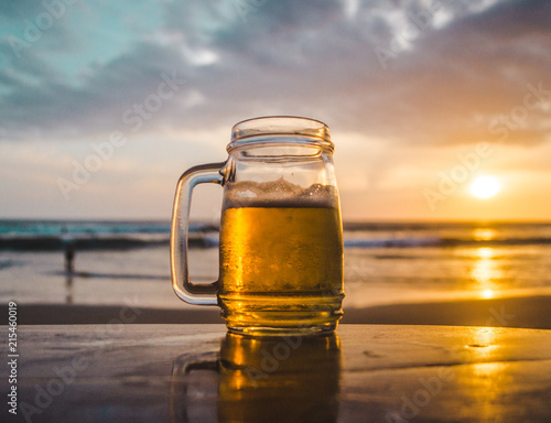 Glass jar of beer on a wooden table looking out at the ocean during sunset