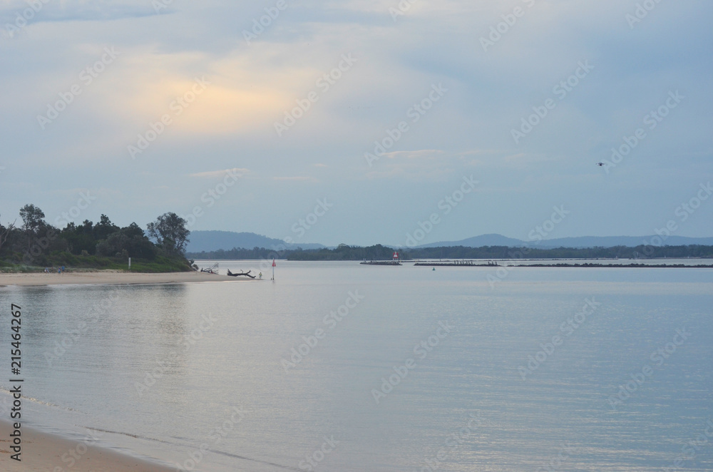 Late afternoon over a river inlet where it joins the ocean. A man is fishing from the beach. The sky is overcast.
