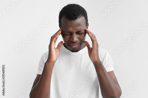 Young African American dark skin man in blank white t shirt isolated on gray background, feeling pain, touching temples with hands, feeling uneasy, tired and discontent