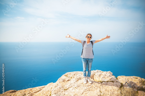 Young beautiful woman tourist girl standing on rocks over blue sea, with open hands, summer travel and vacations