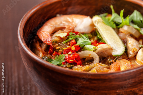 Asian Oriental Cuisine. Japanese spicy soup with seafood, shrimps, vegetables, lime, parsley, and hot pepper, in a plate of red clay, stands on a table in a restaurant. Copy space, selective focus