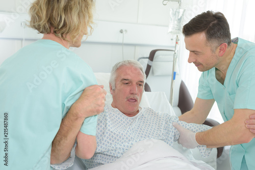 older man lying in hospital bed with nurses helping him