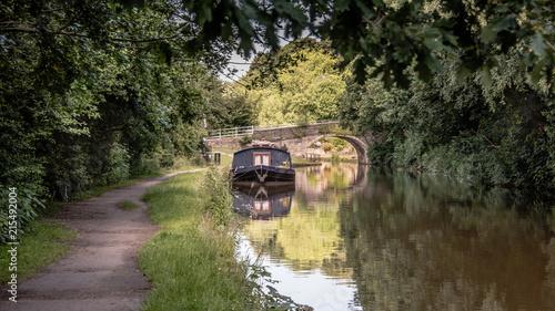 A peaceful scene looking down a canal towpath. There is a narrow boat moored up in front of an old road bridge crossing the water.. The scene is set in between trees at the side of the canal