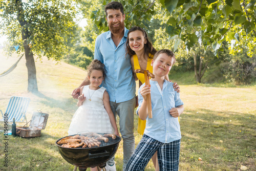 Portrait of a happy family with two cute children, a boy and a girl, looking at camera while standing outdoors near the barbecue grill on a green hill in summer