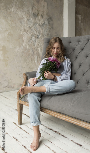 Blonde beautiful model with curly hair sitting on the grey sofa. Girl looking on the pink flowers.