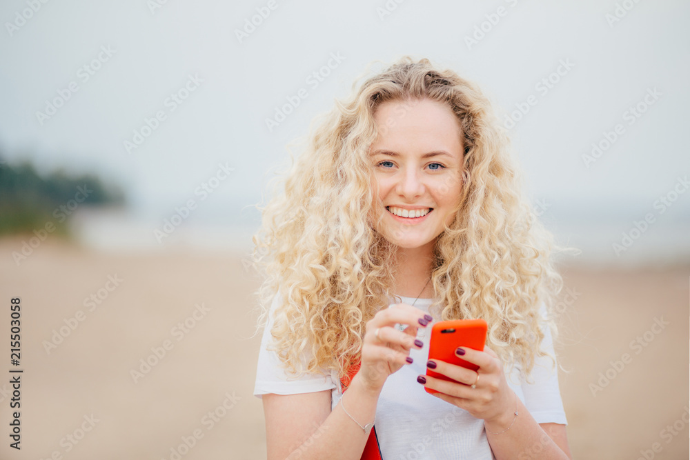 Horizontal shot of good looking young blonde female has curly hair, holds orange smart phone, messages with friends, poses against blurred beach background, rests in tropical country with lover