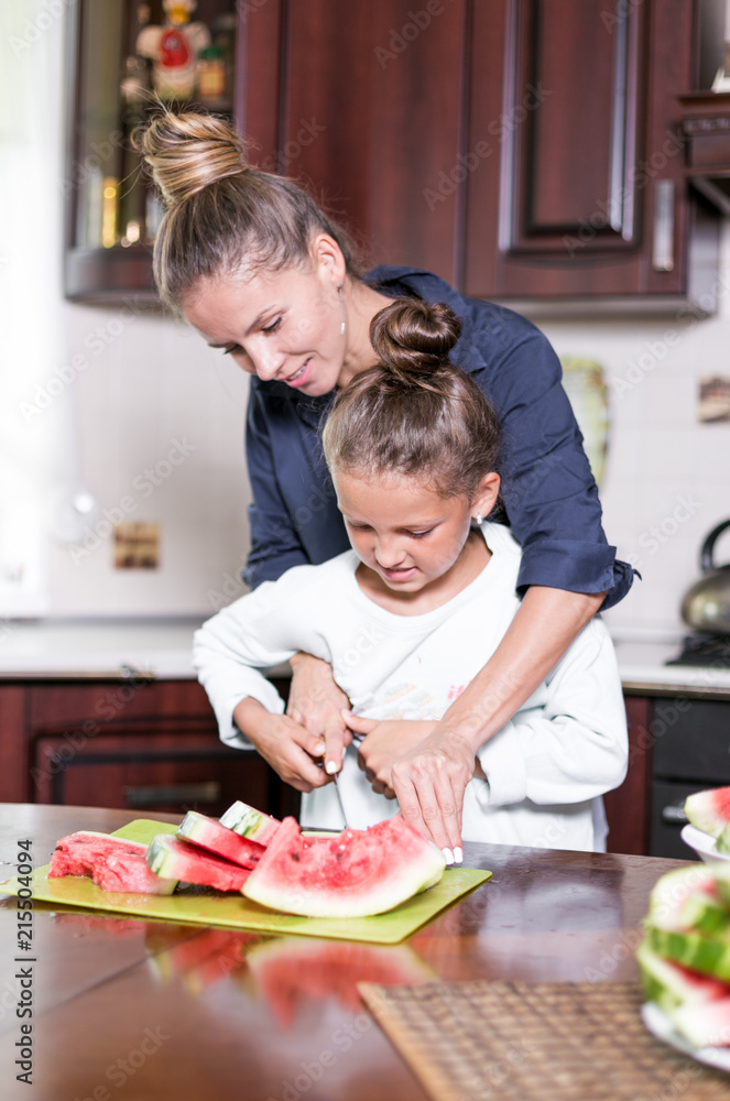 Cute little girl and her beautiful mom are cutting fruits, red watermelon and smiling while cooking in kitchen at home. Happy Family