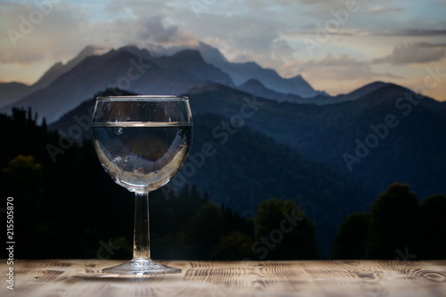 A glass glass glass with clean water stands on a wooden table against a mountain landscape.