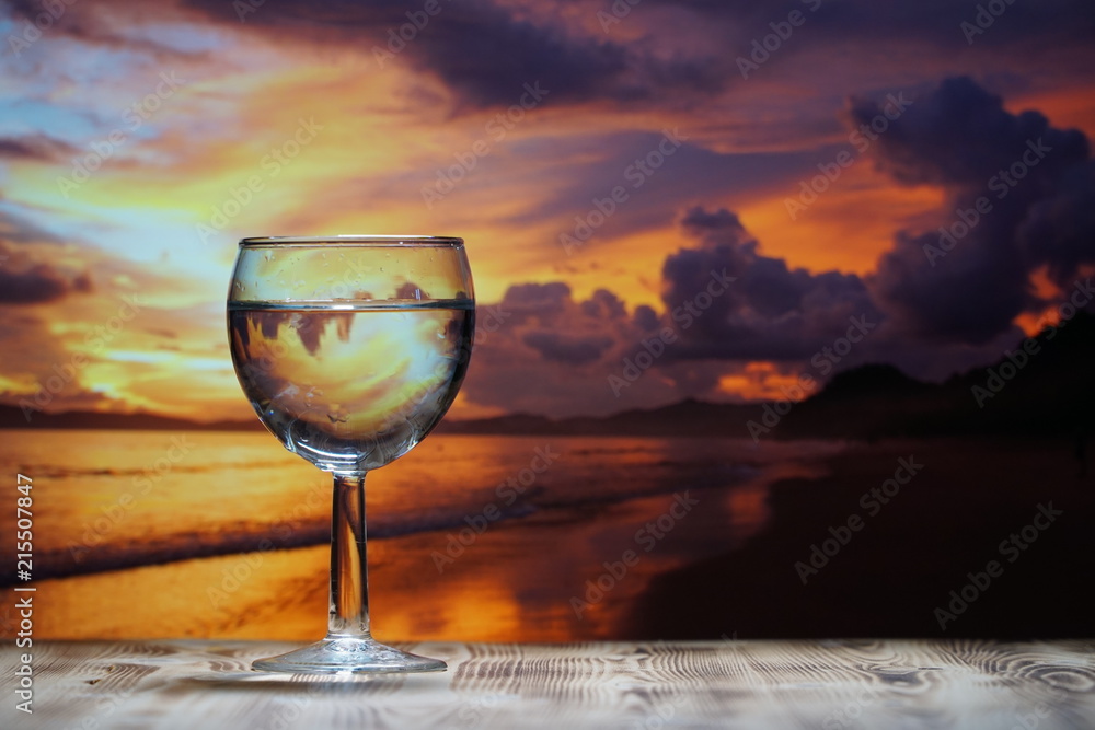 A glass of drinking water is on a wooden table against the background of the sea sunset.
