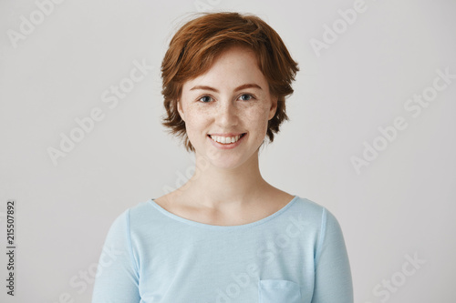 Friendly cute redhead european female colleague with freckles, smiling broadly, standing with confident and happy expression against gray background, being in family circle on weekend