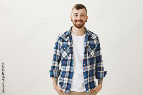 Good-looing confident adult boyfriend with flesh-tunnels and beard standing in blue plaid shirt over gray background, smiling friendly and joyfully, holding hands in pockets